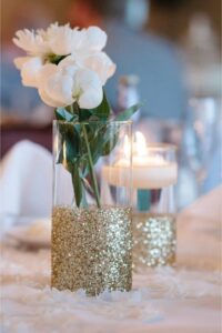 Glass vase with gold glitter with flower and tea light used as DIY wedding decorations on table.