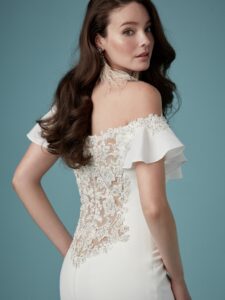 A sheath wedding gown with Chardon crepe, an illusion back with shimmering beaded floral lace motifs, an off-the shoulder neckline, soft ruffle sleeves accented with beaded floral lace motifs, crystal buttons, and a zipper closure.